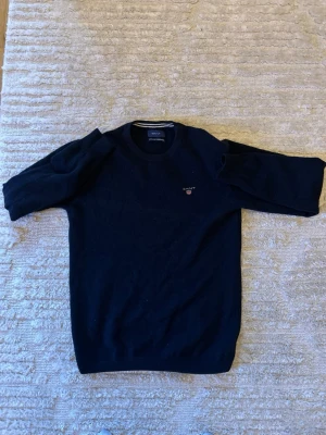 Mörkblå stickad tröja från Gant - Klassisk mörkblå stickad tröja från Gant med rund halsringning och diskret broderad logga på bröstet. Tröjan har långa ärmar och ribbade muddar vid ärmslut och nederkant. Perfekt för lager på lager och stilrena outfits.