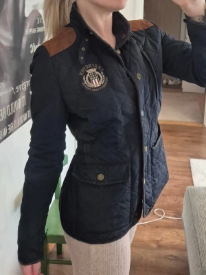 Mörkblå quiltad jacka med bruna detaljer - Snygg mörkblå quiltad jacka från Lexington med bruna axeldetaljer i mocka, tryckknappar och två stora fickor framtill. Jackan har broderad logga på bröstet och markerad midja för en schysst passform. Perfekt för höst och vår.