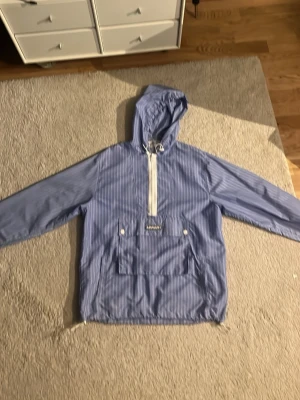 Blåvit randig vindjacka Axel Arigato - Snygg blå vindjacka från Axel Arigato med vita tunna ränder och vit dragkedja framtill. Jackan har huva med dragsko, stor magficka och justerbar nederkant. Perfekt för blåsiga dagar och enkel att matcha med olika outfits.