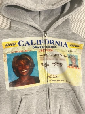 Grå hoodie med WH ID-tryck - Säljer en grå zip-hoodie med stor och print av ett California körkort på framsidan. Hoodien har huva med dragsko, fickor framtill och ribbade muddar. Materialet är mjukt och bekvämt, perfekt för en avslappnad streetwear-look.