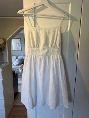 Vit kort sommarklänning med snörning - Söt vit kort klänning med tunna axelband och smockad midja. Klänningen har en öppen rygg med snörningsdetalj och är tillverkad i ett lätt, strukturerat tyg 