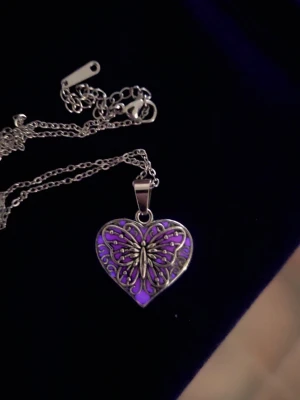 Halsband med hjärthänge och fjäril - Snyggt halsband i silverfärgat rostfritt stål med ett hjärtformat hänge. Hänget har en detaljrik fjäril i metall ovanpå en bakgrund som skiftar mellan lila och rosa beroende på ljus. Kedjan är tunn och elegant, perfekt för att ge din outfit en unik touch. Vanligt dagsljus utifrån aktiverar effekten och hänger ger ifrån sig ett svagt lila sken i mörker. Kedjan är 45 cm och hänget ca 2 cm.