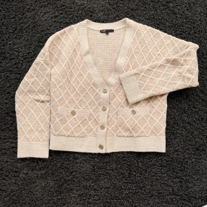 Beige kofta från Maje med rutmönster - Snygg beige kofta från Maje med diskret rutmönster och v-ringad hals. Koftan har två framfickor med dekorativa knappar i champagnefärg och ribbade muddar. Perfekt för lager på lager och ger en chic touch till din outfit.