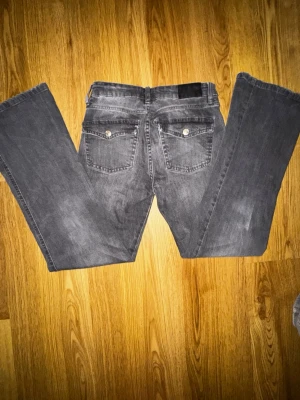 Grå bootcut jeans från lager 157 - Säljer ett par gråa bootcut jeans från H&M med klassisk femficksdesign och snygga bakfickor med lock och knapp. Jeansen har en lätt tvättad look och är tillverkade i bomull med lite stretch för skön passform.
