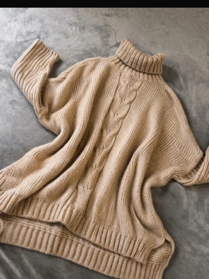 Beige stickad polotröja med flätmönster - Mysig beige stickad polotröja med bred ribbad polokrage och dekorativt flätmönster framtill. Tröjan har avslappnad passform, långa ärmar och ribbade muddar vid ärmslut och nederkant. Perfekt för kalla dagar och enkel att styla till olika outfits.