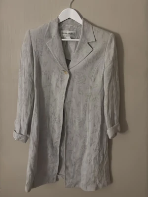 Ljusgrå kavaj från Pierre Cardin - Stilren ljusgrå kavaj från Pierre Cardin med diskret mönster i tyget. Lång modell med klassisk krage och en knapp framtill. Tillverkad i Italien, materialet har en lätt glansig finish och känns lyxigt. Perfekt för dig som vill ha en unik och elegant look.