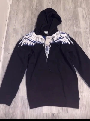 Svart hoodie med vingeprint från Marcelo Burlon - Säljer en svart hoodie från Marcelo Burlon med ikoniskt vitt och blått vingeprint över axlar och bröst. Hoodien har huva, långa ärmar och ribbade muddar. Perfekt för dig som vill sticka ut med en unik design.