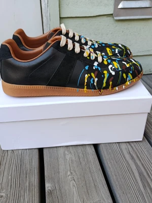 Maison margiela  - Unika svarta sneakers med coola färgstänk i blått, gult, vitt och brunt på ovandelen. Skorna har bruna sulor och beige snören, samt en insida i brunt läderimitation. Perfekta för dig som vill sticka ut med din stil och gillar konstnärliga detaljer.