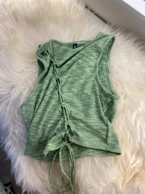 Grön ribbad linnetopp med snörning - Trendig grön linnetopp med ribbad struktur och snörning framtill. Toppen är ärmlös och har en lätt croppad passform, perfekt för att styla med höga jeans eller kjol. Snygg detalj med knyt längst ner och diskret rutigt mönster.