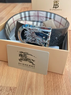 Burberry rutigt bälte med silverdetaljer - Snyggt bälte från Burberry med klassiskt rutigt mönster i beige, svart och rött. Bältet har en exklusiv silverfärgad spänne med Burberrys ikoniska riddare. Perfekt accessoar för att lyfta din outfit.