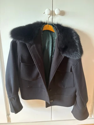 Svart pälsjacka från Meotine XS/S - Snygg svart pälsjacka från Meotine med stor fluffig fuskpäls runt kragen. Jackan har två stora fickor framtill, knäppning med knappar och en rak, lite boxig passform. Fodrad med mörkgrönt tyg för extra komfort och stil. Säljs med defekt, flertal knappar som lossnat men går att fixa om man är bra på det. Nypris ca 3000kr