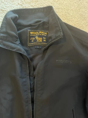 Svart bomberjacka från Woolrich - Snygg svart bomberjacka från Woolrich med diskret logga på bröstet och dragkedja framtill. Jackan har två fickor med dragkedja och ribbade muddar vid ärmslut och midja. Perfekt för dig som vill ha en stilren och klassisk look.