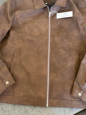 Brun skinnjacka från Eva Jonsson - Snygg brun skinnjacka från Eva Jonsson med rak passform, klassisk krage och silverfärgad dragkedja. Jackan har två sidofickor och tryckknappar vid ärmsluten. Perfekt för dig som vill ha en stilren och tidlös look. Den är väldigt tunn. 