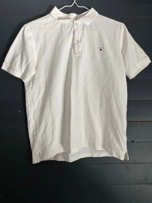 Vit pikétröja från Gant - Klassisk vit pikétröja från Gant med liten broderad logga på bröstet. Tröjan har korta ärmar, krage och knappar framtill. Tillverkad i mjuk bomull som känns skön mot huden. Perfekt till jeans eller shorts för en clean look.