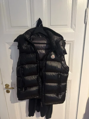 Svart dunväst från Moncler med luva - Snygg svart dunväst från Moncler med luva och två dragkedjefickor framtill. Västen har en glansig finish och klassisk Moncler-logga på bröstet. Perfekt för lager-på-lager och håller dig varm under kyliga dagar.