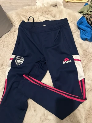 Arsenal träningsbyxor från Adidas - Mörkblå träningsbyxor från Adidas med Arsenal-logga på ena benet och Adidas-logga på andra. Byxorna har vita och rosa detaljer längs sidorna samt dragkedja vid benslut. Tillverkade i ett lätt och stretchigt material, perfekta för träning eller chill. Liten i storlek 