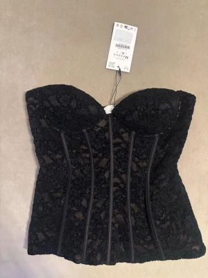 Svart spetskorsett från Zara - Trendig svart korsett från Zara med vacker spets och markerade sömmar framtill. Axelbandslös modell med vadderade kupor och nude-färgad insida. Perfekt för att skapa en snygg siluett och ge outfiten en extra edge.