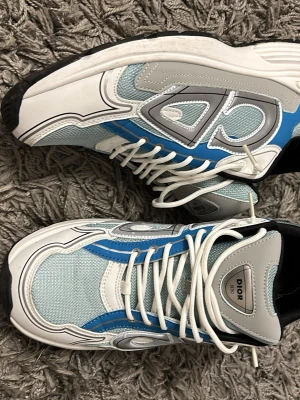  sneakers blå och grå - Säljer ett par  sneakers i blått, grått och vitt med coola reflekterande detaljer och meshpaneler. Skorna har vita snören, svart sula på sidan. Perfekta för dig som gillar streetwear och exklusiva märken.