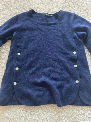 Mörkblå stickad tröja från Ellos - Mysig mörkblå stickad tröja från Ellos med rund halsringning och dekorativa vita knappar på sidorna. Tröjan har lång ärm och en mjuk, lite fluffig struktur. Perfekt för lager på lager och enkel att matcha med jeans eller kjol.