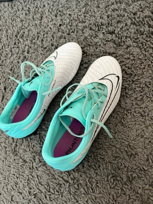 Nike Phantom GT2 vita fotbollsskor - Säljer ett par Nike Phantom GT2 fotbollsskor i vitt med turkosa detaljer och snörning. Skorna har en stor svart Nike-logga på sidan och lila innersula. Ovandelen är tillverkad i syntetmaterial för bra bollkontroll och passform. Perfekta för dig som vill sticka ut på planen.