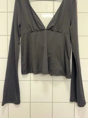 Mörkbrun omlottopp med knyt - Trendig mörkbrun omlottopp med djup v-ringning och långa ärmar. Toppen har ett snyggt knyt i ryggen som ger en unik detalj. Tillverkad i mjukt och stretchigt material som sitter bekvämt på kroppen. Perfekt för dig som gillar stilrena plagg med twist.