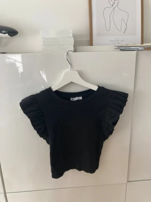 Svart topp med volangärmar från Zara - Snygg svart topp från Zara med ribbad struktur och volangdetaljer på ärmarna. Modellen är ärmlös och har en rund halsringning. Perfekt för dig som vill ha en enkel men ändå trendig look.