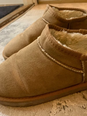 Uggs - Mysiga bruna tofflor från 157 i mockaimitation med mjukt, vitt foder i fårskinnslook. Tofflorna har rundad tå och låg ankel, perfekta för att hålla fötterna varma. Klassisk design med synliga sömmar och rejäl sula.