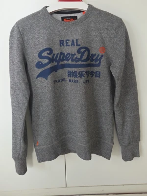 Grå sweatshirt från Superdry - Snygg grå sweatshirt från Superdry med blå logga och japansk text på bröstet. Tröjan har rund halsringning, långa ärmar och ribbade muddar vid ärmslut och nederkant. Perfekt för en avslappnad och trendig stil.