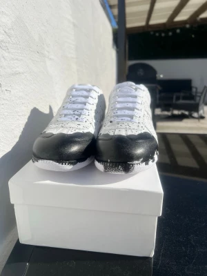 Maison margielas med färg  - Unika vita sneakers med svart färgstänk och svart detalj framtill. Skorna har lågt skaft, vita skosnören och är tillverkade i slätt lädermaterial. Perfekta för dig som vill sticka ut med en kreativ och modern look.