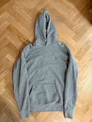 Grå hoodie från GANT med huva - Snygg grå hoodie från GANT med klassisk huva och dragsko. Hoodien har en liten GANT-logga broderad på bröstet och en stor magficka framtill. Tillverkad i mjukt bomullsmaterial som är skönt att ha på sig. Perfekt för en chill och avslappnad stil.