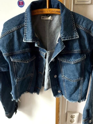 Cropped blå jeansjacka Perfect Jeans - Säljer en croppad blå jeansjacka från Perfect Jeans. Jackan har klassisk krage, två bröstfickor med lock och knappar framtill. Rå kant nedtill för en edgy look. Perfekt för dig som vill ha en trendig och avslappnad stil.