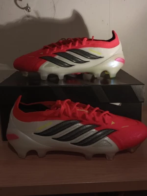 Adidas Predator elite F G fotbollsskor röd/vit HELT NYA!!! - HELT NYA. Säljer ett par Adidas Predator ELITE FG  fotbollsskor i röd, vit och svart färg med svarta och silverfärgade detaljer. Skorna har klassisk snörning, låg profil och mönstrad sula för bra grepp på planen. Tillverkade i syntetmaterial och levereras med originalpåse och original låda.Till pojkar män och killar