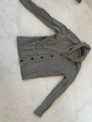 Grå kofta från Polo Ralph Lauren - Snygg grå kofta med långa ärmar, knäppning framtill med svarta knappar och två praktiska fickor. Koftan har en enkel och stilren design, perfekt att slänga på sig över en t-shirt. Materialet ser ut att vara mjukt och bekvämt, troligen bomull eller bomullsblandning. Passar även M