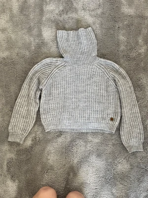 Grå polotröja stickad från Fjällräven - Säljer en grå ribbstickad polotröja från Fjällräven. Tröjan har hög krage, långa ärmar och en liten läderdetalj med logga vid nederkanten. Perfekt för kalla dagar och riktigt mysig att bära.