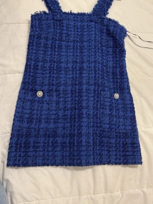 Blå kort klänning med fickor - Säljer en blå kort klänning från Monki i strukturerat tyg med breda axelband och två fickor framtill med dekorativa knappar. Klänningen har en rutig textur och är perfekt för dig som gillar en trendig och ungdomlig stil.