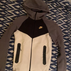 Nike tech fleece, XS - Nike hoodie i färgblock med svart bröstparti, grå ärmar och huva samt vit front. Guldig Nike-logga på bröstet, hel dragkedja och två fickor med dragkedja. Perfekt för chill eller träning, riktigt snygg och sportig vibe. Nästan aldrig använd, kan finnas katthår på den tyvärr.