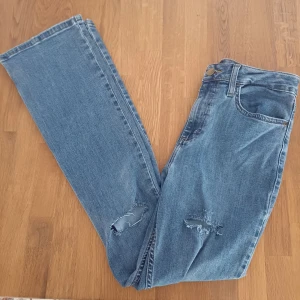 Blå mom jeans med slitningar - Säljer ett par blå mom jeans med trendiga slitningar på båda benen. Jeansen har hög midja, raka ben och klassisk femficksdesign. 