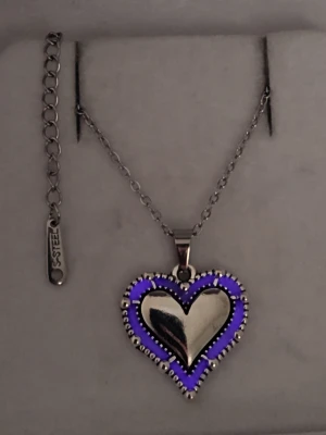 Halsband med hjärthänge i stål - Lyser lila i mörker! - Snyggt halsband i rostfritt stål med ett hjärthänge. Hänget har en blank, silverfärgad yta och är dekorerat med små pärlliknande detaljer runt kanten och lyser lila i mörker efter att ha varit i vanligt dagsljus eller solljus en kort stund. Bär det som ett vanligt halsband så sköter sig effekten själv. Lyser i flera timmar i mörker, både hänget o kedjan tål vatten, lång livslängd. Perfekt accessoar för en trendig look. Handgjort.