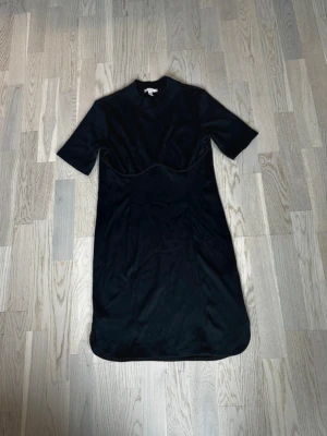 Svart kortärmad klänning - En stilren svart klänning med korta ärmar och rund hals. Klänningen har en lätt markerad söm över midjan och rundad nederkant med små slitsar i sidorna. Materialet ser ut att vara mjukt och följsamt, perfekt för en enkel och clean look. Rök och djurfritt hem. Pruta gärna !🌸