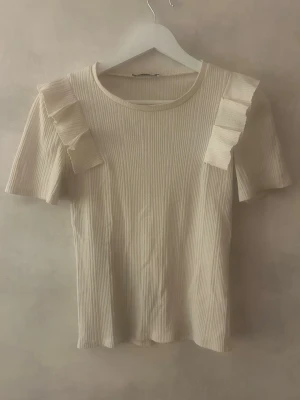 Ribbad cremefärgad topp med volang - Supersöt ribbad topp i cremevitt med korta ärmar och volangdetaljer på axlarna. Perfekt till jeans eller kjol för en trendig look. Mjuk och stretchig känsla, rund halsringning och smickrande passform.