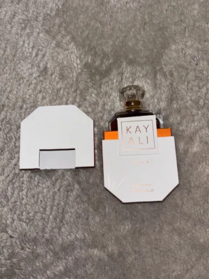 Kayali Vanilla 28 Eau de Parfum - Kayali Vanilla 28 Eau de Parfum i en elegant, genomskinlig glasflaska med guld- och vitt detaljer. Flaskan har en unik, kantig form och levereras i en vit, geometrisk förpackning med guldtext. Doften är känd för sina varma, söta vaniljtoner.