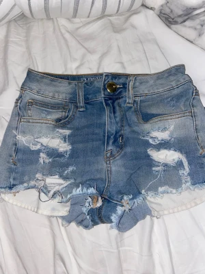 Blå slitna jeansshorts American Eagle - Säljer ett par blå jeansshorts från American Eagle Outfitters med slitna detaljer och råa kanter. Shortsen har klassisk femficksdesign, knappstängning och är tillverkade i stretchigt denim. Perfekta för en avslappnad och trendig look.