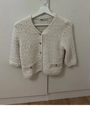Beige stickad kofta från Zara - Snygg beige stickad kofta från Zara med v-ringning och dekorativa knappar framtill. Koftan har två små fickor med knappdetaljer och ett grovt stickat mönster som ger en trendig look. Perfekt att slänga över en topp för extra stilpoäng.