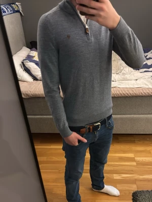Grå halvzip-tröja från Morris - Snygg blå/grå stickad tröja från Morris med halv dragkedja och diskret logga broderad på bröstet. Tillverkad i 100% merinoull för en mjuk och skön känsla. Perfekt till jeans eller chinos för en clean look. Sitter lite tajtare än M.