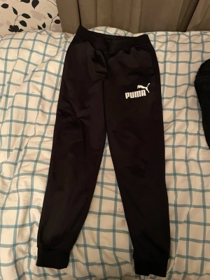 Svarta mjukisbyxor från Puma - Snygga svarta mjukisbyxor från Puma med vit logga på låret. Byxorna har resår i midjan och vid bensluten för en skön passform. Perfekta för chill eller träning. Tillverkade i ett mjukt och följsamt material.
