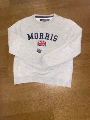 Grå sweatshirt från Morris - Snygg ljusgrå sweatshirt från Morris med mörkblå text och brittisk flagga broderad på bröstet. Klassisk rund halsringning och långa ärmar. Perfekt för en avslappnad och stilren look.