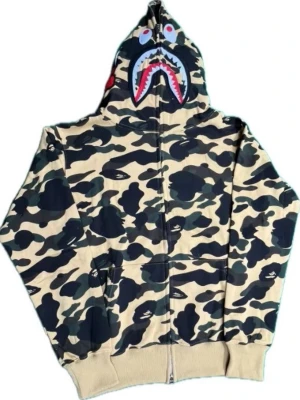 BAPE camo zipper - Säljer en ikonisk BAPE hoodie i beige och grönt camomönster med dragkedja och stor huva. Huvan har det klassiska hajmotivet med tänder och ögon, samt broderade detaljer på sidan. Hoodien har ribbade muddar och känguruficka framtill. Perfekt för streetwear-stilen.