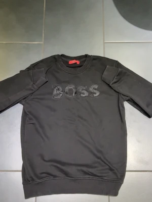 Svart sweatshirt från BOSS - Säljer en svart sweatshirt från BOSS med broderad logga på bröstet. Tröjan har rund halsringning, långa ärmar och ribbade muddar vid ärmslut och nederkant. Perfekt för en avslappnad och stilren look.