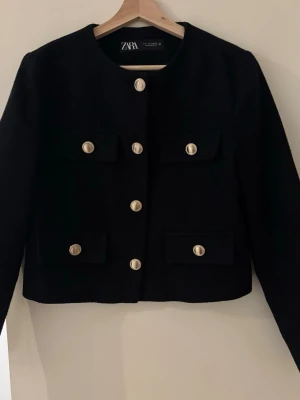 Svart kavaj med guldknappar från Zara - Stilren svart kavaj från Zara med rund halsringning och sex dekorativa guldknappar framtill. Perfekt för en trendig och elegant look. Inga defekter!