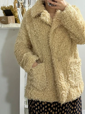 Beige fluffig teddyjacka - Supermysig teddyjacka i beige med stora fickor framtill och bred krage. Jackan har en oversized passform och är gjord i ett lurvigt, mjukt material som ger en trendig look. Perfekt för kalla dagar när du vill vara både varm och stylish.